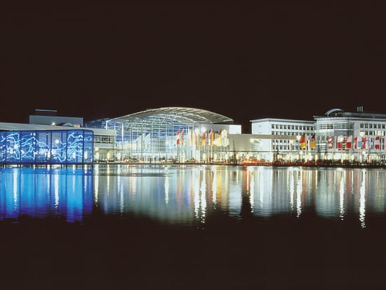 Beleuchtete Gebäude der Messe München spiegeln sich nachts im Wasser.