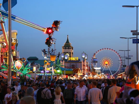 Das Münchner Oktoberfest in der Abenddämmerung, mit beleuchteten Fahrgeschäften, Riesenrad und einer belebten Menschenmenge.