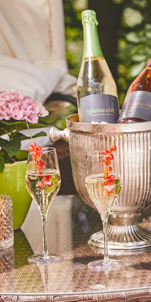 Stylishly arranged champagne cooler with two Freizzante bottles, decorated sparkling wine glasses and snacks on a lounge table on the summery hotel terrace.