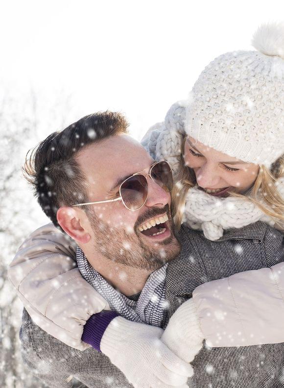 Lachendes Paar in Winterkleidung beim ausgelassenen Spaziergang im Schnee - der Mann trägt die Frau lachend auf dem Rücken.