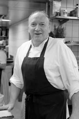 Lächelnder Koch in weißer Kochjacke und schwarzer Schürze in der Restaurantküche.
