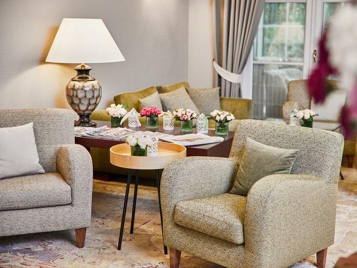 Im Vordergrund steht ein kleiner Holztisch zwischen zwei Sesseln in beige und pastellgrün bezogen, dahinter ein Sideboard mit Zeitschriften und einer angeschalteten Lampe vor einer pastellgrünen Couch mit Dekokissen.
