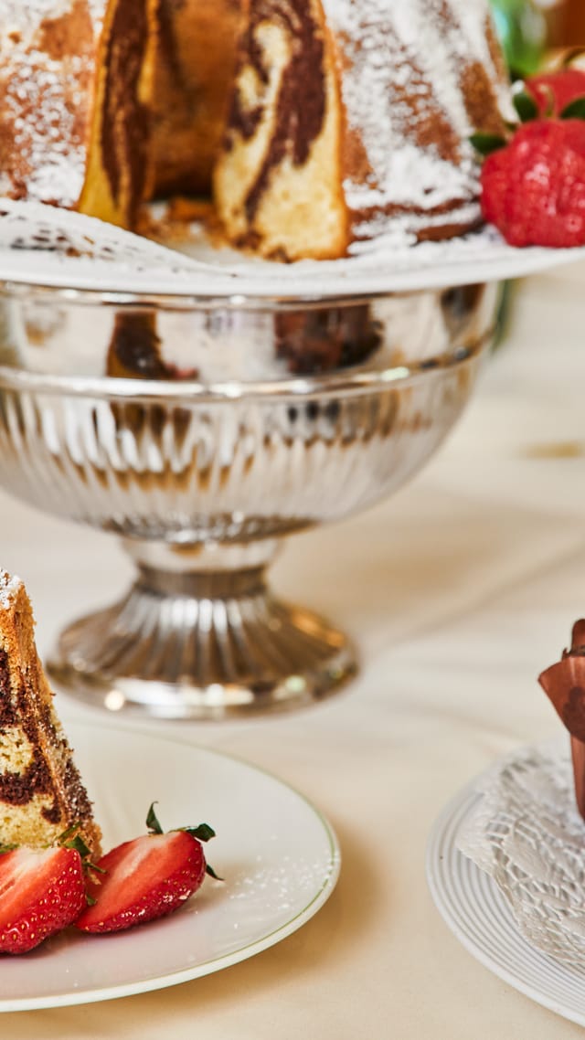 Marmorkuchen mit Puderzucker und Erdbeeren auf einem silbernen Tortenständer, davor ein serviertes Stück, daneben Schokomuffins.