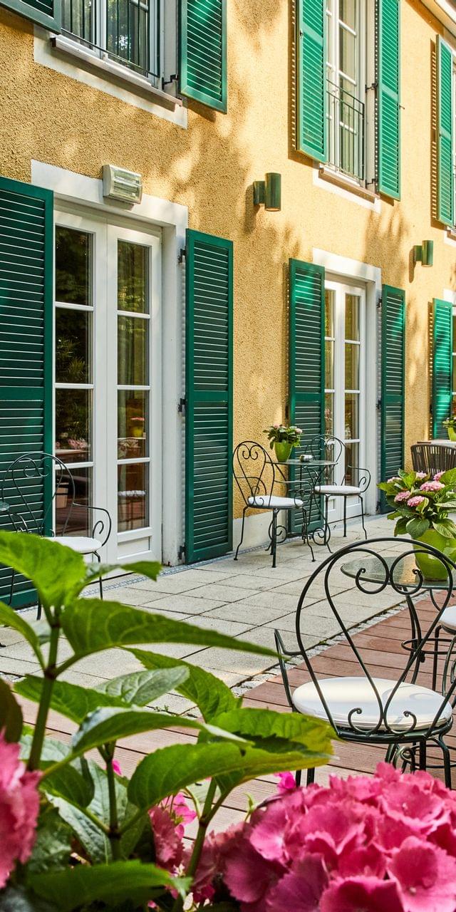 Weitläufige Hotelterrasse mit mehreren Sitzgruppen aus Schmiedeeisen und Holz, üppig blühenden Hortensien und der charakteristischen gelben Fassade mit grünen Fensterläden.