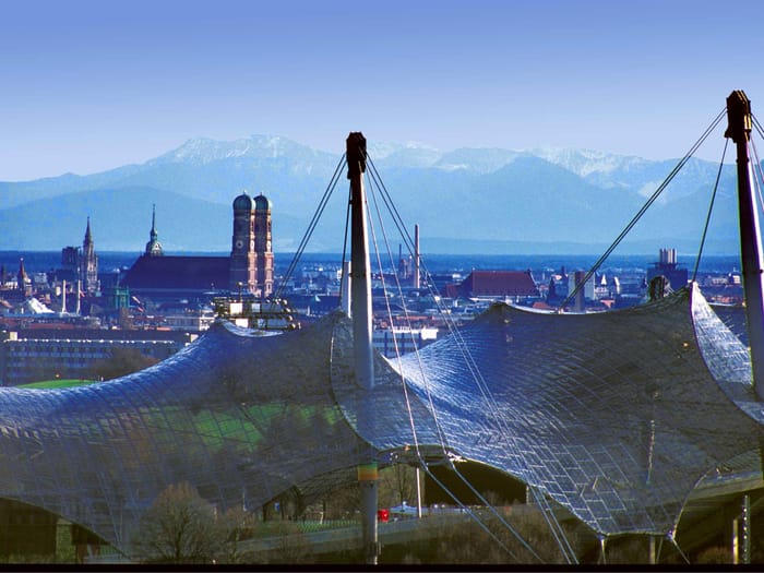 Blick über München mit Frauenkirche und schneebedeckten Alpen im Hintergrund, im Vordergrund Masten des Olympiaparks.