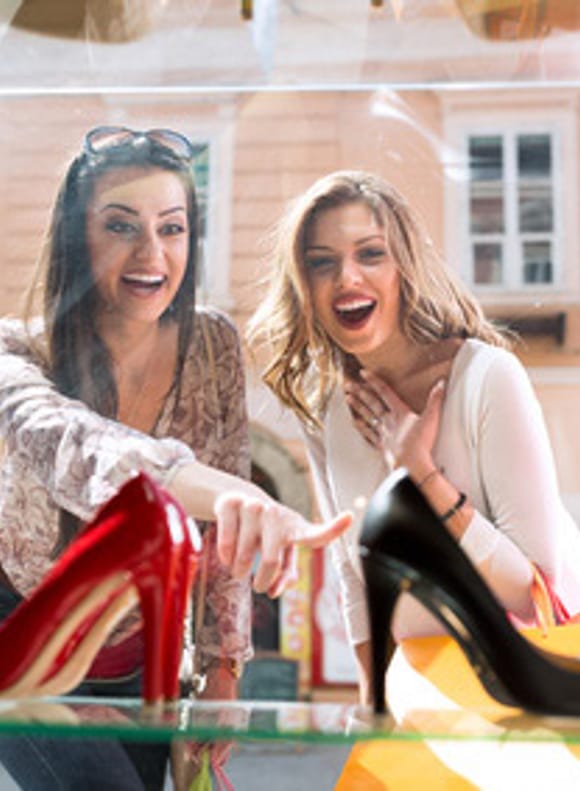 Two excited women gazing in amazement at a shop window displaying colourful shoes – shopping fun and city trip vibes in downtown Munich.