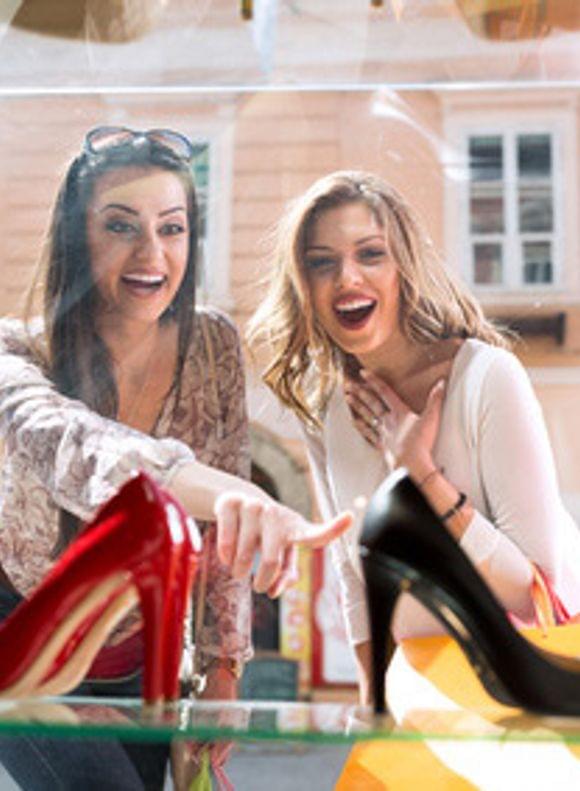 Two excited women gazing in amazement at a shop window displaying colourful shoes – shopping fun and city trip vibes in downtown Munich.