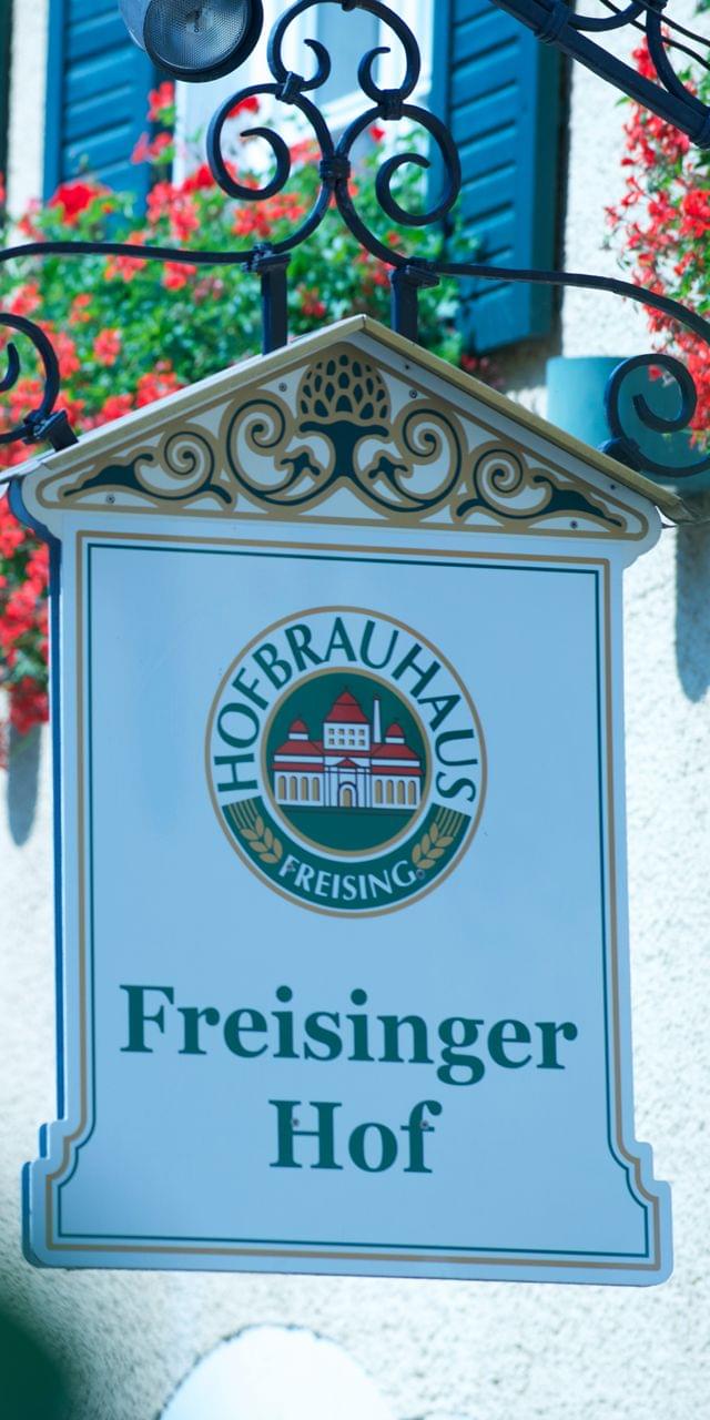 Zoom auf das Schild Freisinger Hof über der Eingangstür des Restaurants mit roten Geranien in den Blumenkästen daneben.