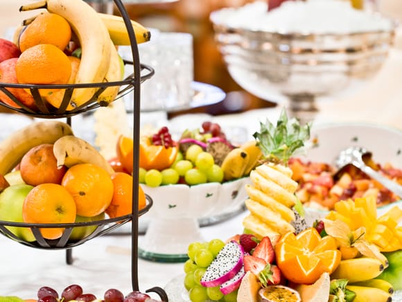 Reichhaltige Obst-Station mit Etagere voller Bananen, Orangen und Äpfel, umgeben von kunstvoll angerichteten Tellern mit geschnittener Ananas, Drachenfrucht, Kiwi, Grapefruit und weiteren Früchten.