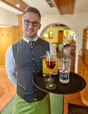 Service staff member in Bavarian traditional costume serving a tray with aperitif, orange juice and water in the restaurant.
