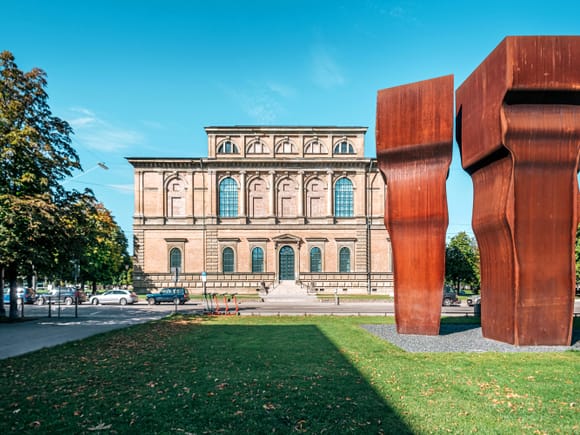 Die Alte Pinakothek in München mit ihrer klassizistischen Fassade, davor eine markante moderne Skulptur aus Cortenstahl auf grüner Wiese.