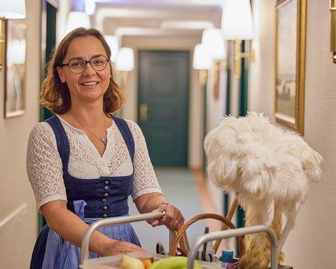 Freundlich lächelnde Hausdame in Dirndl schiebt einen Servicewagen durch den elegant beleuchteten Hotelflur.