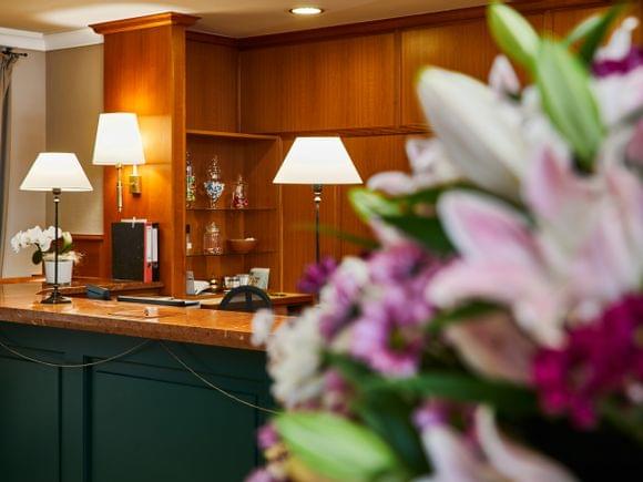 Blick durch einen unscharfen Lilien-Blumenstrauß auf die warme Hotelrezeption des Freisinger Hof München: Marmortheke mit petrolgrüner Front, Holzvertäfelung, klassischen Tischlampen, einer weißen Orchidee auf dem Tresen und gläsernen Bonbonièren im Regaleinbau.
