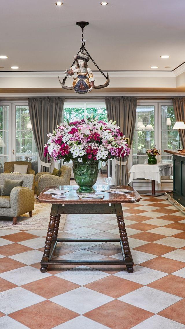 Erster Blick durch die Eingangstür in die klassische Hotellobby: Ein prächtiges Blumengesteck auf einem Antiktisch empfängt die Gäste, flankiert von einer eleganten Rezeption und einem gemütlichen Sitzbereich – verbunden durch einen charakteristischen Schachbrettboden.