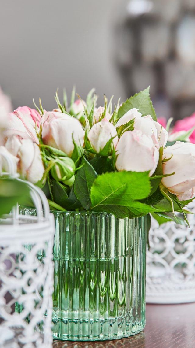 Kleine Blumenvasen aus grünem Glas mit rosa und pinken blumen und weißen kleinen Vogelkäfigen dazwischen in einer Reihe.