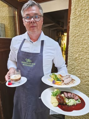 Service employee of the Freisinger Hof restaurant carrying two festively presented main courses and a dessert out of the kitchen.