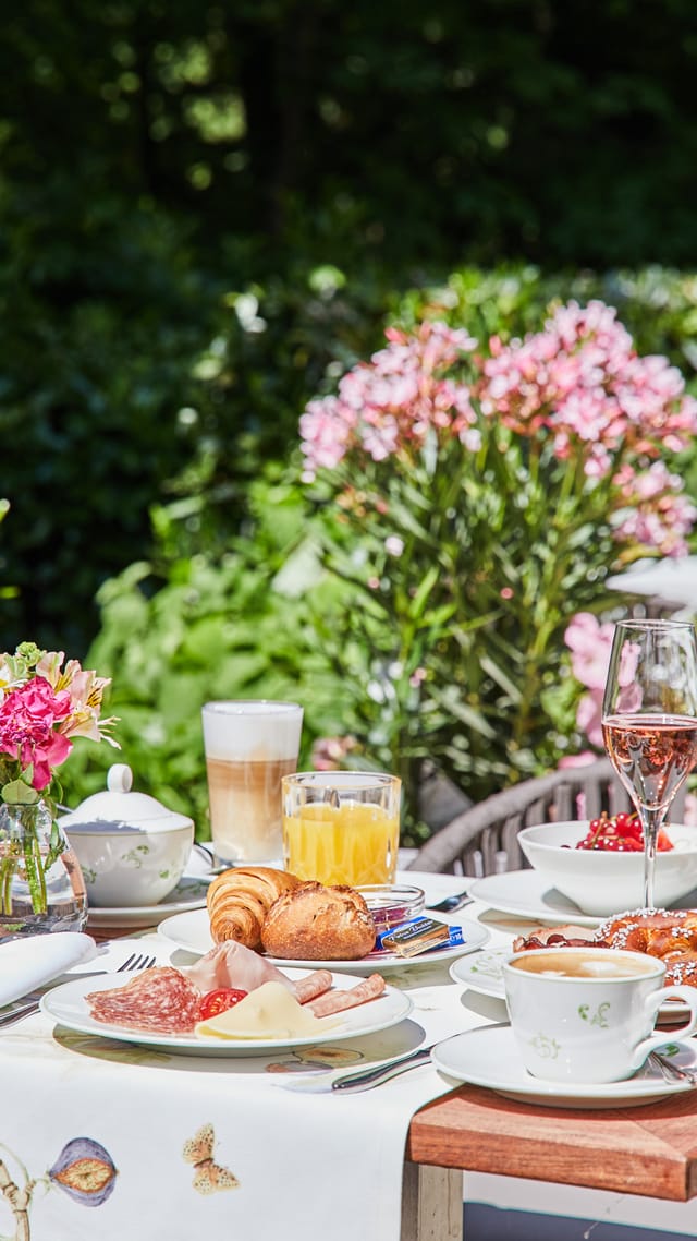 Sonnenbeschienener Terrassentisch mit Gläsern Sekt, Latte Macchiato, frischen Blumen, Croissant, Wurst und Käse, umgeben von Rattanstühlen und blühendem Oleander.