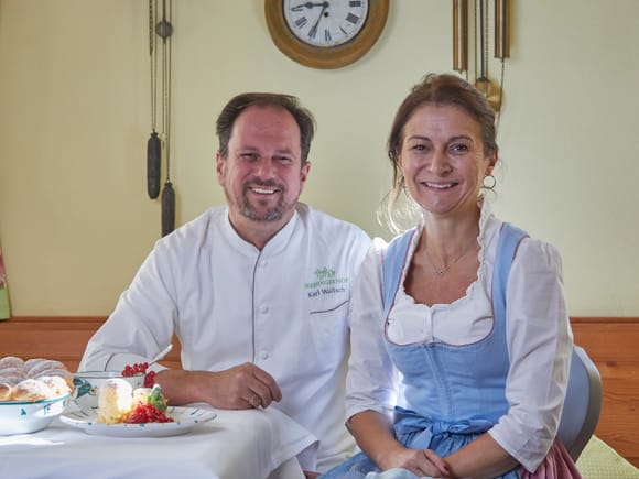 Die Gastgeber des Freisinger Hof München: Küchenchef Karl Wallisch und seine Frau sitzen an einem Tisch mit Buchteln und lächeln in die Kamera. Im Hintergrund sind Wanduhren zu sehen.