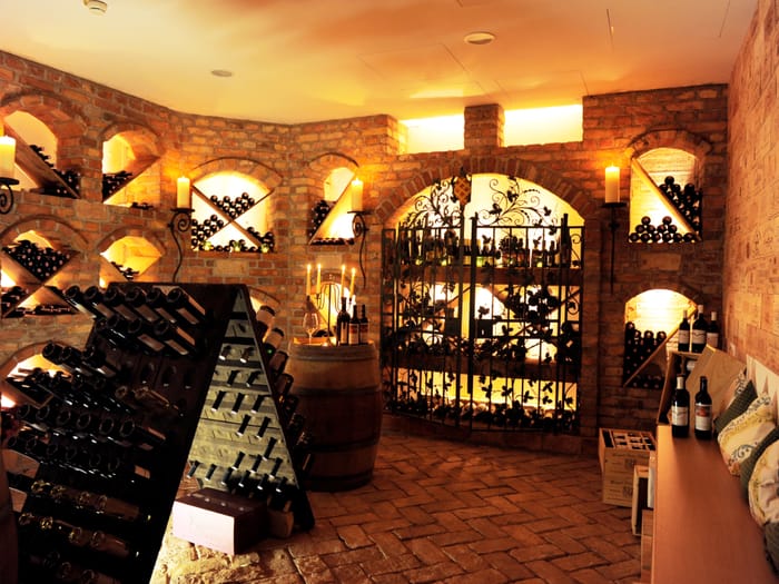 Atmospheric overall view of the historic wine cellar with illuminated diamond racks, wrought-iron vine gate, riddling racks, wooden barrels and a cozy seating nook.