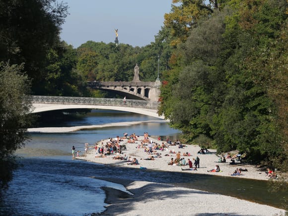 Sonnenbadende auf den Kiesbänken der Isar in München, im Hintergrund die Brücke und die goldene Statue des Friedensengels.