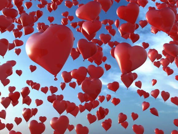 Numerous red heart-shaped balloons rising into the blue sky.