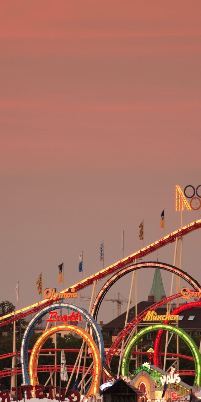 Fahrgeschäft des Münchner Oktoberfests bei Sonnenuntergang, im Hintergrund die Silhouette der Frauenkirche vor rosa-orangefarbenem Himmel.