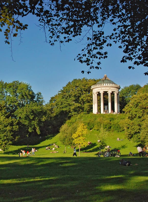 The Monopteros on its hill in Munich's English Garden, framed by tree branches and surrounded by green lawns.