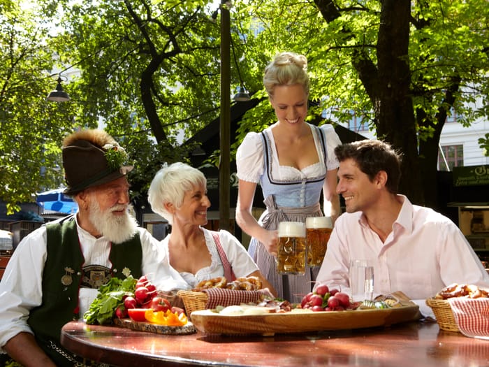 Kellnerin in Tracht bedient Gäste an einem Tisch im schattigen Biergarten.