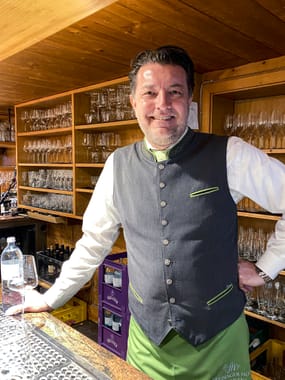 Smiling Freisinger Hof restaurant employee in Bavarian traditional costume leaning relaxed against the bar, with a large selection of wine glasses in the background.
