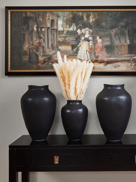 Vier schwarze Vasen mit beigen Ziergräsern auf einer schwarzen Kommode und ein rechteckiges Bild an der Wand.