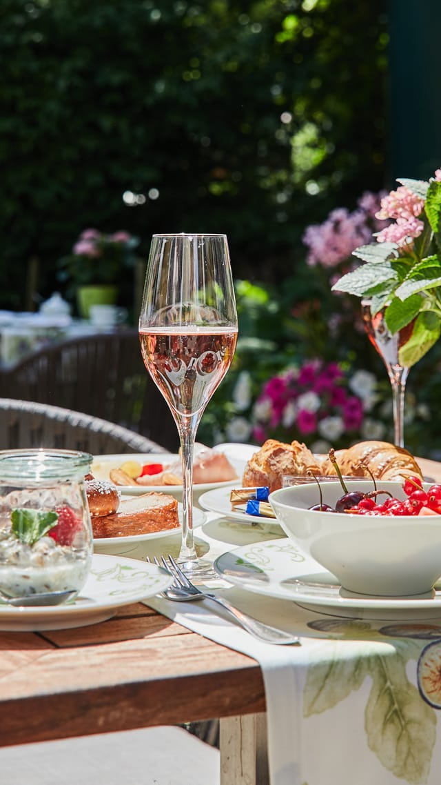 Frühstückstisch im schattigen Garten mit Kirschen, roten Johannisbeeren, Latte Macchiato, Sekt und Infused Water, umgeben von blühenden Hortensien.