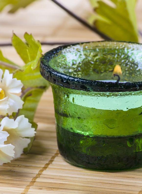 Ein grünes Teelichtglas neben weißen Blüten auf einem Bambusmattentisch – ruhige Wellnessatmosphäre und Entspannung.