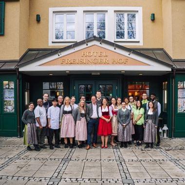 Das Team des Hotel und Restaurant Freisinger Hof posiert gemeinsam vor dem Hoteleingang - viele Mitarbeiterinnen und Mitarbeiter sind in bayerischer Tracht.