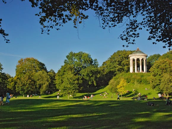 Der Monopteros auf seinem Hügel im Englischen Garten München, gerahmt von Baumzweigen und umgeben von grünen Wiesen.