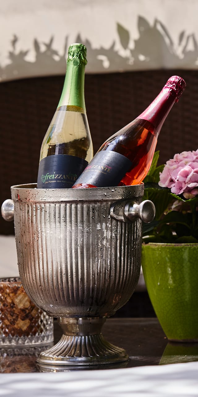 Two bottles of Frizzante in an ice bucket with garnished glasses and a hydrangea on a round lounge table, surrounded by the white cushions of the cozy garden lounge.