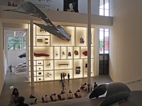 Ausstellungsraum der Pinakothek der Moderne mit Vitrinen voller Design-Exponate und einem Modell, das von der Decke hängt.