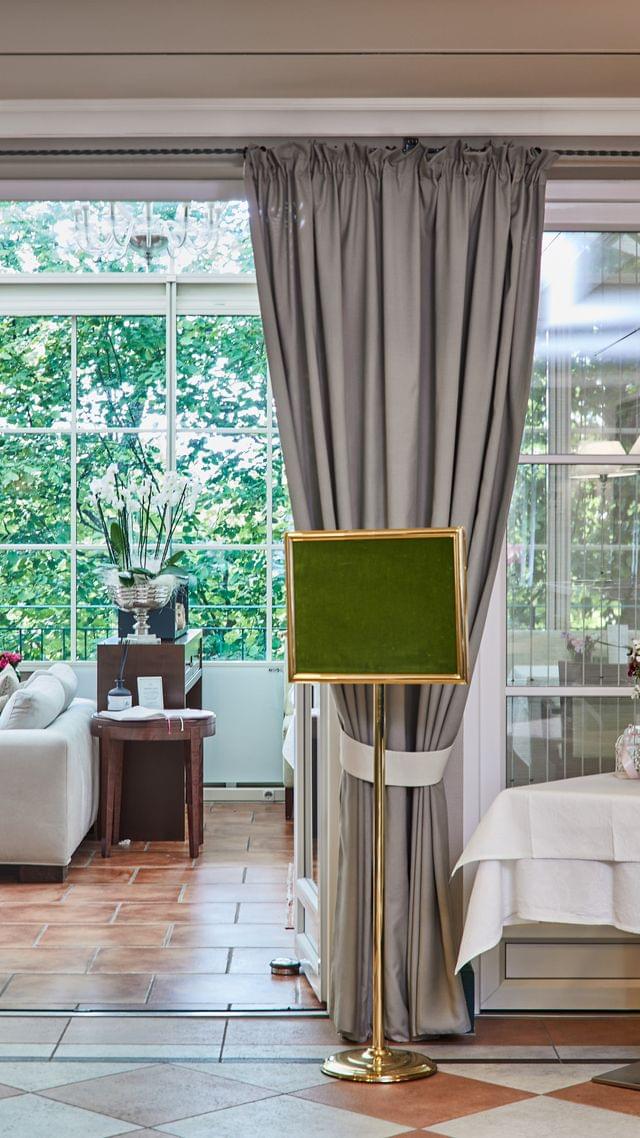 Inviting view through elegant double doors into a light-flooded conservatory — in the foreground a set table with flowers, in the background green trees and a cozy seating area.