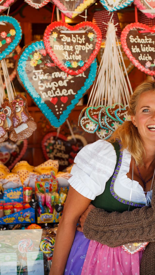 Paar in bayerischer Tracht vor einem Stand mit bunten Lebkuchenherzen auf dem Münchner Oktoberfest.