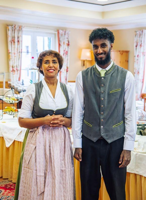 Zwei freundlich lächelnde Servicemitarbeiter in bayerischer Tracht stehen im Frühstücksraum des Hotels.