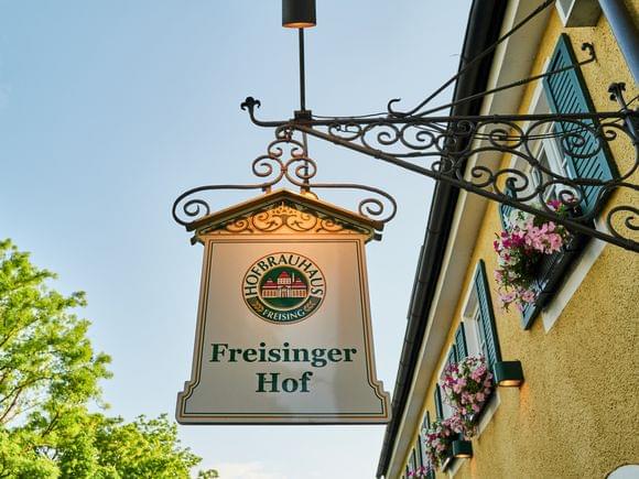 Zoom auf das Schild Freisinger Hof über dem Restauranteingang mit blauem Himmel und leichter Beleuchtung.