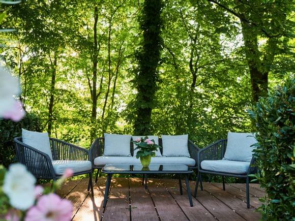 Sitzgruppe vor Laubbäumen auf der Hotelterrasse mit grauen Polstern und Glastisch, auf dem eine kleine Hortensie steht.