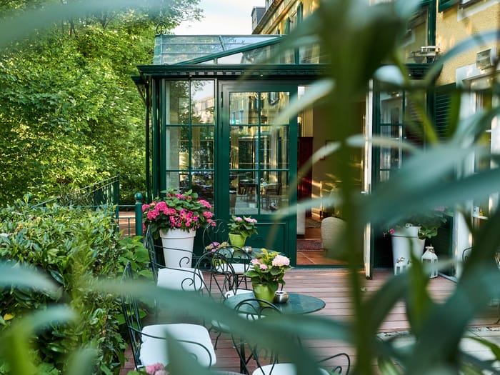 Blick auf den grünen Wintergarten des Hotels mit angrenzender Holzterrasse, schmiedeeisernen Sitzgruppen und pinkfarbenen Hortensien, umgeben von dichtem Grün.