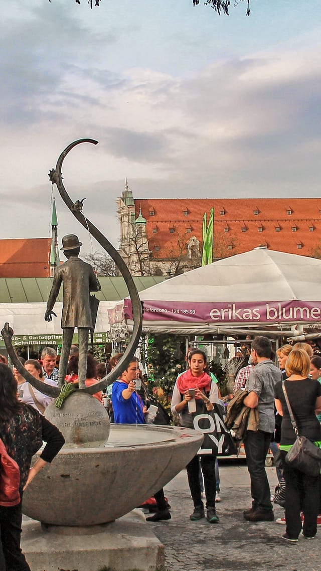 Der Viktualienmarkt München mit einem Brunnen im Vordergrund und dem Turm des Alten Peters im Hintergrund.