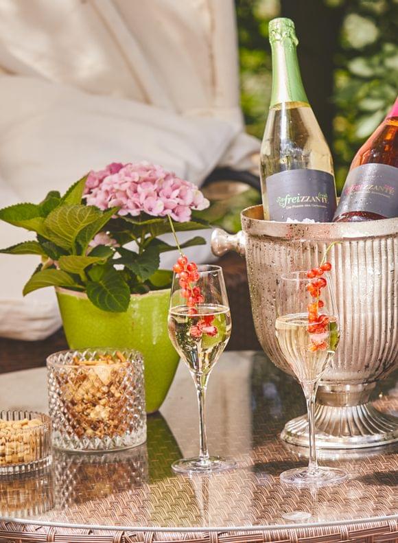 Stilvoll arrangierter Sektkühler mit zwei Freizzante-Flaschen, dekorierten Sektgläsern und Knabbereien auf einem Loungetisch auf der sommerlichen Hotelterrasse.