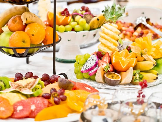 Reichhaltige Obst-Station mit Etagere voller Bananen, Orangen und Äpfel, umgeben von kunstvoll angerichteten Tellern mit geschnittener Ananas, Drachenfrucht, Kiwi, Grapefruit und weiteren Früchten.