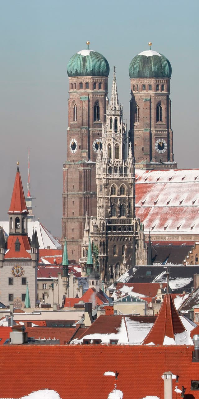 Panoramablick über die schneebedeckten Dächer der Münchner Altstadt mit mehreren Kirchtürmen, darunter der Alte Peter und die Frauenkirche, die die winterliche Skyline prägen.