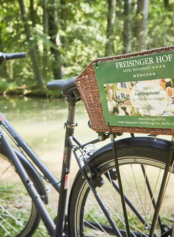 Fahrrad mit Weidenkorb und Werbeschild des Freisinger Hofs „Mein Lieblingshotel am Englischen Garten", abgestellt an einem Bach im Grünen.
