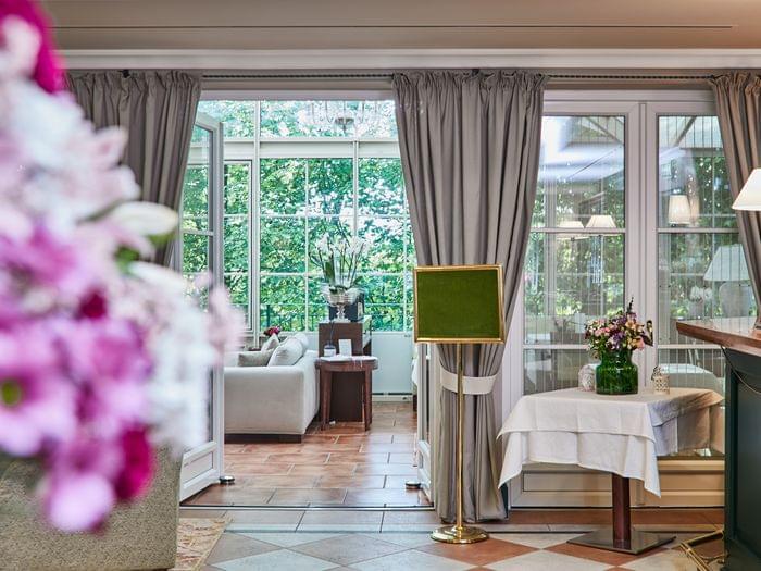 Einladender Durchblick durch elegante Flügeltüren in einen lichtdurchfluteten Wintergarten – im Vordergrund ein gedeckter Tisch mit Blumen, im Hintergrund grüne Bäume und ein gemütlicher Sitzbereich.