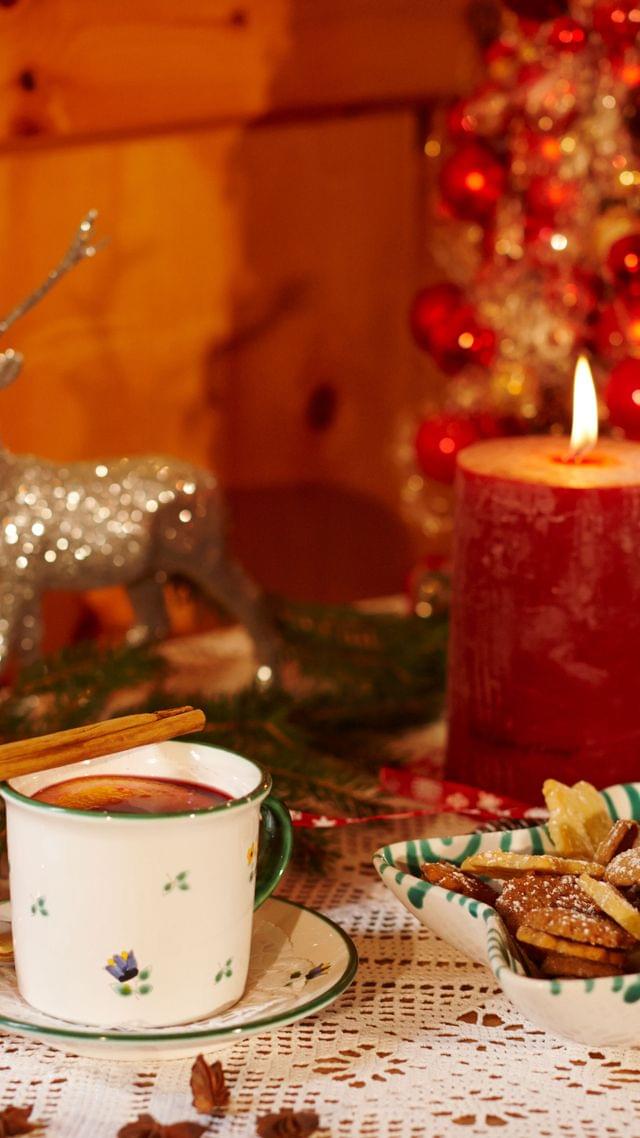 Weihnachtsdekoration in gold-rot Tönen, eine rote Kerze in der Mitte, dahinter ein Weihnachtsbaum aus kleinen Christbaumkugeln, eine glitzernde Hirschfigur und eine Tasse Zimtkakao davor.