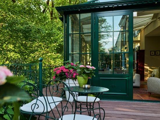 Peaceful seating corner on the hotel terrace with wrought iron table, hydrangeas and a view into the green conservatory — surrounded by old trees.